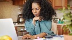 Young black woman sitting in kitchen at the table with papers scattered and laptop open